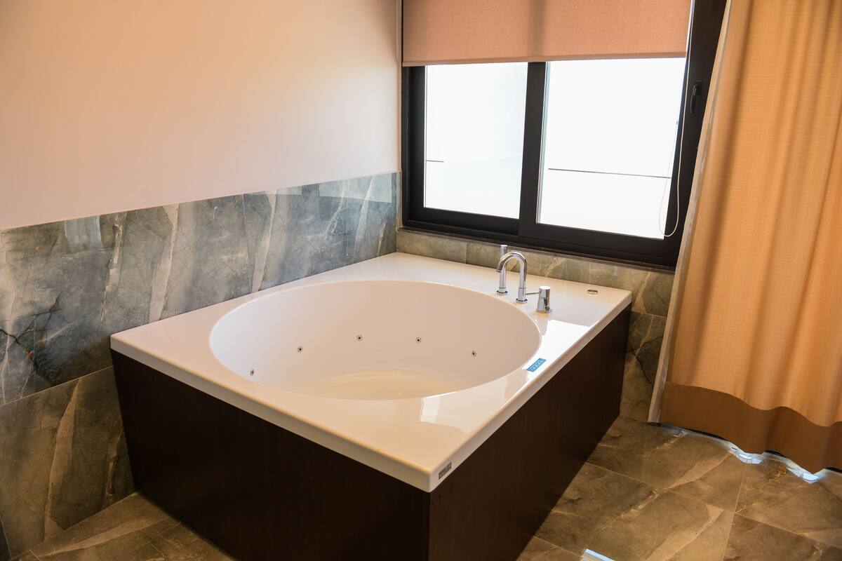 A modern bathroom with a freestanding bathtub, a window with blinds, and stone-tiled flooring.