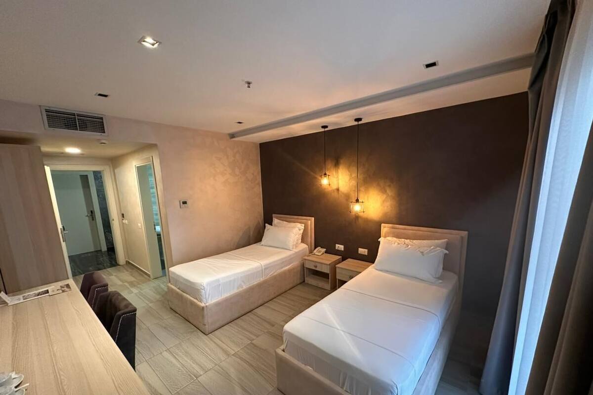 A hotel room with two beds, a desk, a chair, a bedside table, a lamp, and a door leading to a bathroom.