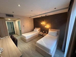 A hotel room with two beds, a desk, a chair, a bedside table, a lamp, and a door leading to a bathroom. - Brilliant Hotel & Spa (Golem)