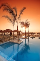 Zona junto a la piscina con palmeras, sillas de descanso y sombrillas al atardecer.