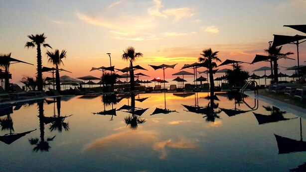 A resort pool with palm trees and umbrellas, reflecting the sunset. - Brilliant Hotel & Spa (Golem)