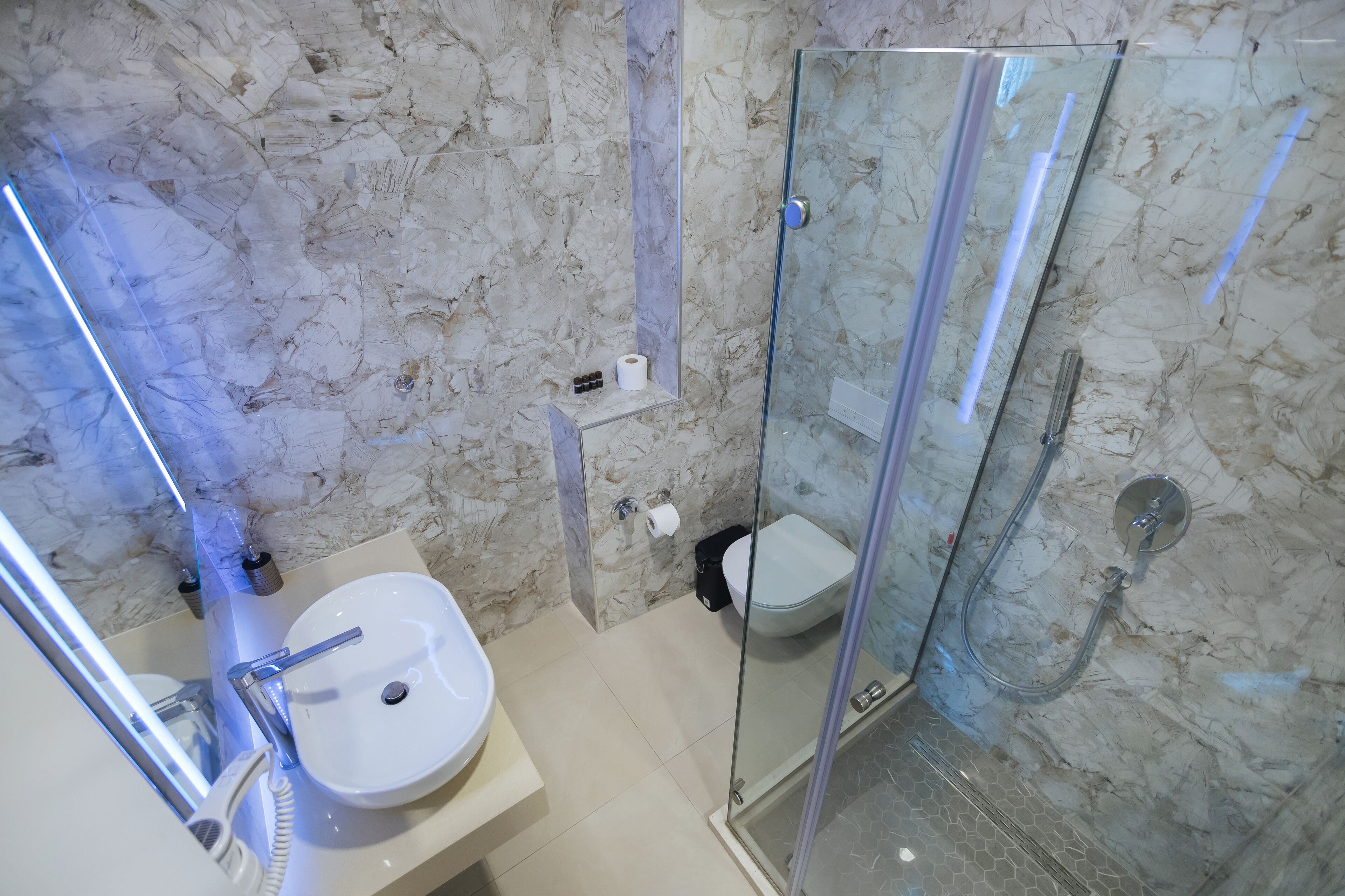 A modern bathroom with a marble wall, a glass-enclosed shower, a white sink, and a toilet.