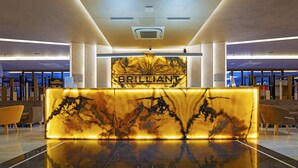 A modern lobby with a marble reception desk, a chandelier, and a view of the city through large windows. - Brilliant Hotel & Spa (Golem)