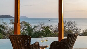 Une vue sur la mer depuis un espace extérieur aménagé avec des chaises et une table en osier, d’où l’on aperçoit la mer et une île au loin.