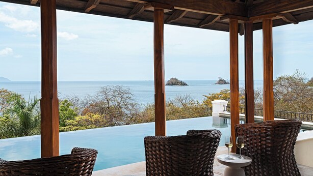 Un espace extérieur aménagé, abrité et doté de mobilier en osier, offrant une vue sur une piscine et la mer.
