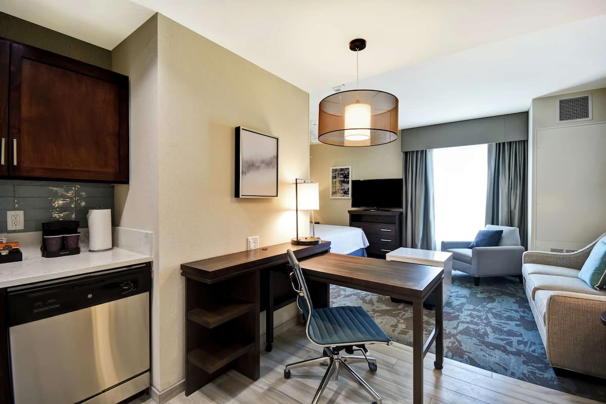 A hotel room with a kitchenette, a dining area with a table and chairs, a sofa, a TV, and a large window with curtains.