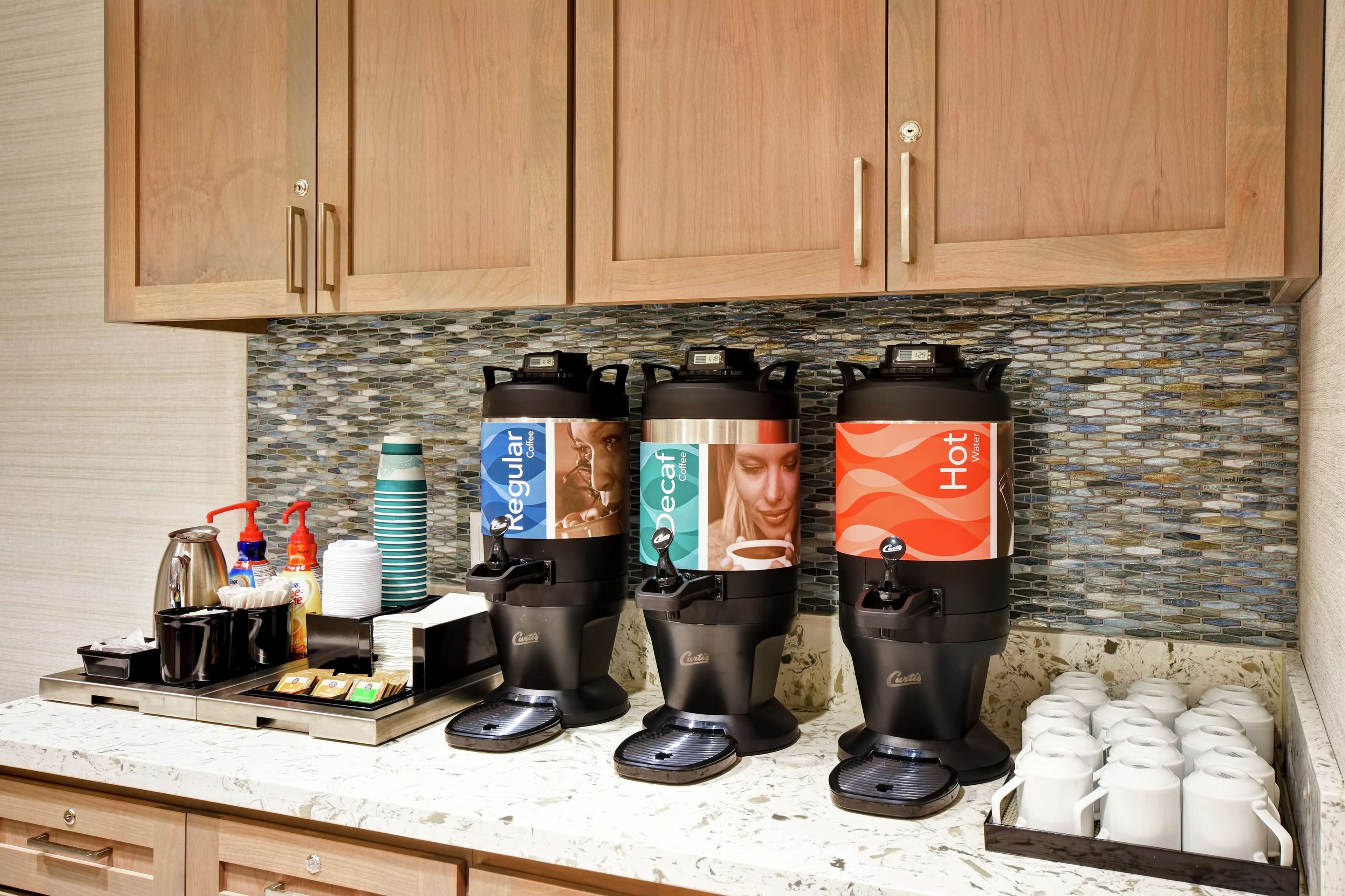 A coffee station with three dispensers labeled Regular, Decaf, and Hot, and a tray with coffee cups and condiments.