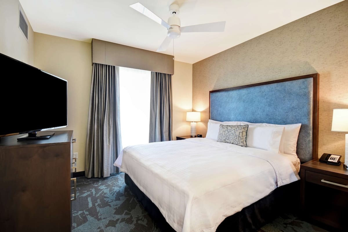 A hotel room with a large bed, a flat-screen TV, a ceiling fan, and a nightstand with a lamp.