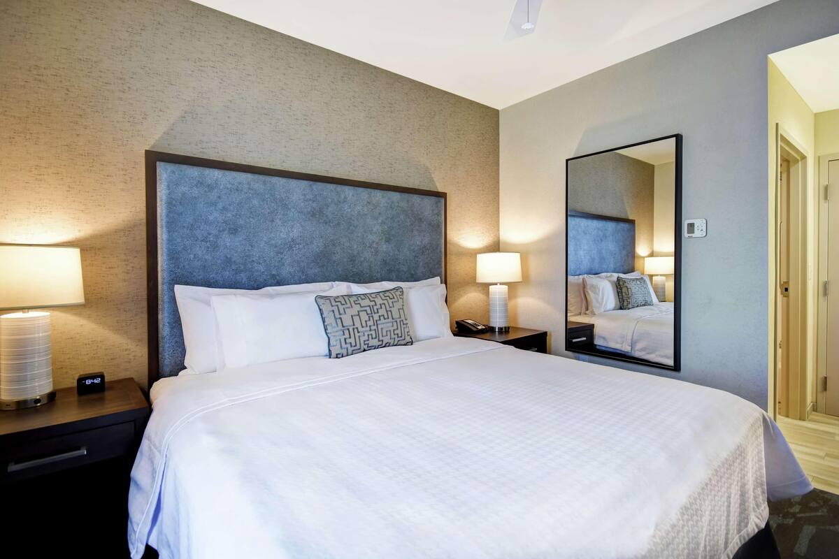 Une chambre d’hôtel avec un grand lit, deux tables de chevet avec des lampes, une commode, un miroir et une porte donnant sur une autre pièce.