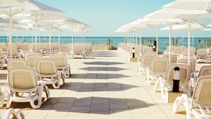 Un área de descanso en la playa con hileras de sillones blancos y sombrillas, orientada al océano.