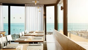 Un restaurante con vistas al mar, con mesas de madera y sillas blancas, ventanas amplias y lámparas colgantes.