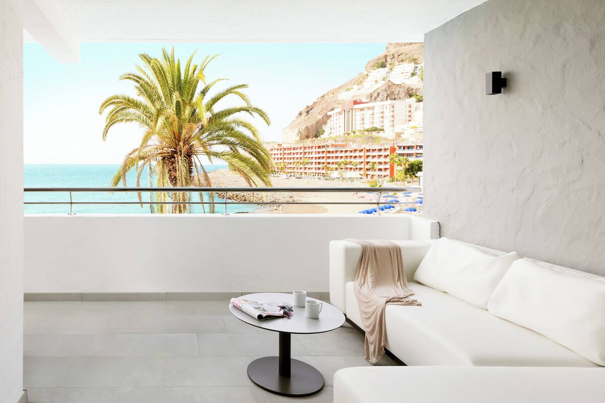 A modern living room with a white sofa, a small round table with a magazine, and a view of the beach and hotel buildings.