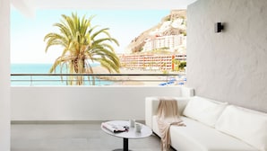 Una sala de estar moderna con un sofá blanco, una pequeña mesa redonda con una revista y vistas a la playa y edificios de hotel.
