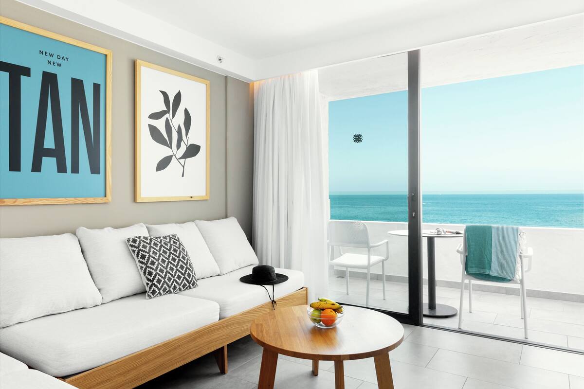 A modern living room with a sofa, a round wooden table with a fruit bowl, and a balcony with a view of the sea.