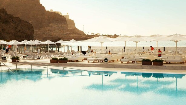 Zona de piscina con sillones de descanso y sombrillas, una piscina cristalina y un acantilado rocoso al fondo.