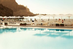 A poolside area with lounge chairs and umbrellas, a clear pool, and a rocky cliff in the background. - Ocean Beach Club Gran Canaria (Mogan)