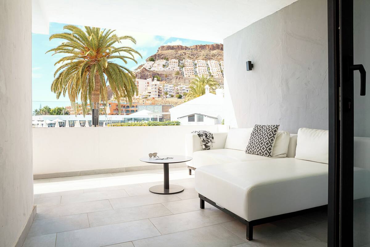 A modern living room with a white sofa, a small round table, and a large window offering a view of a palm tree and buildings.