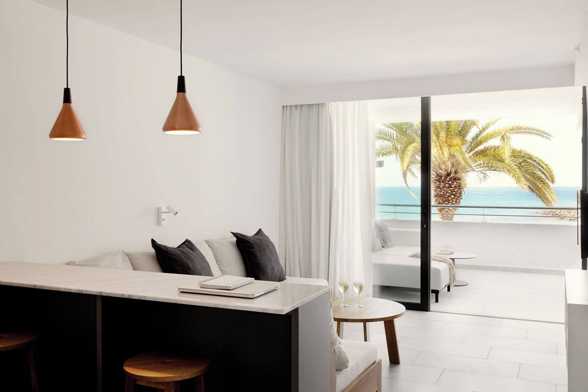 A modern hotel room with a balcony, a white sofa, a small table, and a bar area.