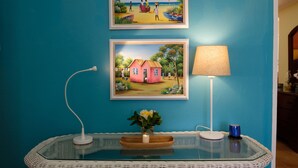 A room with a teal wall, a glass table with a white charger, a lamp, and two framed paintings depicting tropical scenes. - Dreamin Villa  Villa 4 (Duncans)