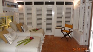 A bedroom with a bed, a chair, a shelf with books, and a door with louvered shutters. - Dreamin Villa  Villa 4 (Duncans)