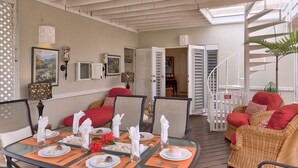 A dining area with a round table set for four, surrounded by wicker chairs with red cushions. A staircase with a white railing leads to an upper level. - Dreamin Villa  Villa 4 (Duncans)