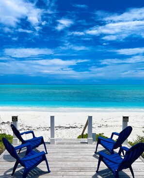 Uitzicht op een strand met twee blauwe stoelen op een houten terras, een bord met het verbod op open vuur, en een bord met een lijst van voorzieningen zoals een restaurant, spa en strandactiviteiten.