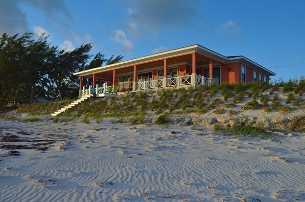 Een huis aan het strand met een overdekte veranda, een trap naar de ingang en een zandstrand op de voorgrond.