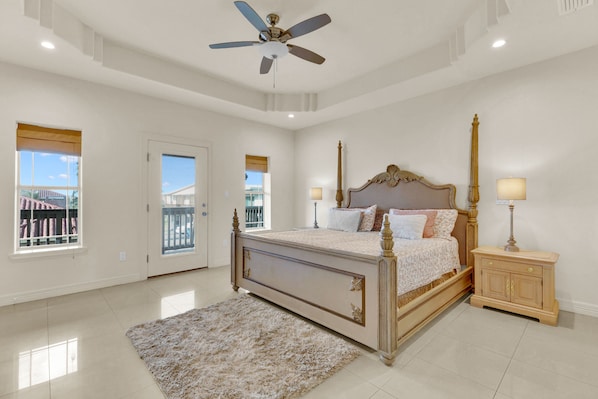 A bedroom with a large bed, a ceiling fan, a nightstand, and a view of the outside.