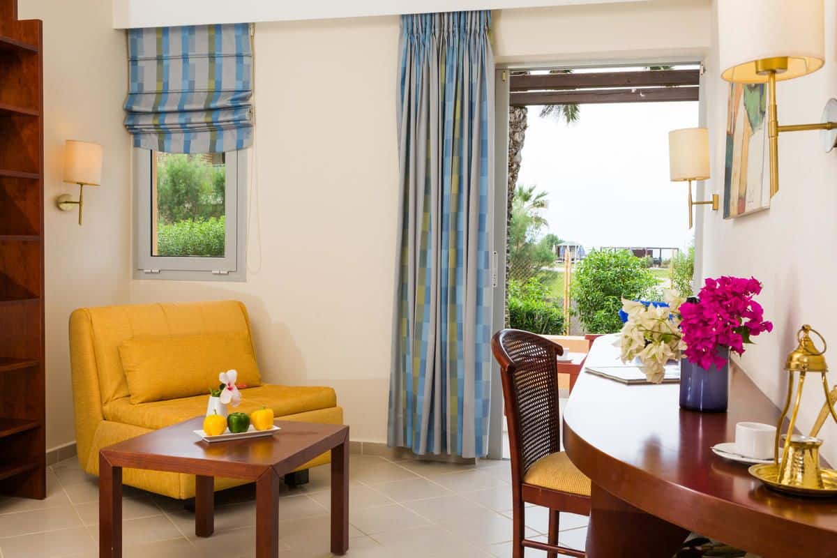 A room with a yellow sofa, a wooden table with a tray of fruit, a wicker chair, a desk with a vase of flowers, and a view of greenery through a window with blue curtains.