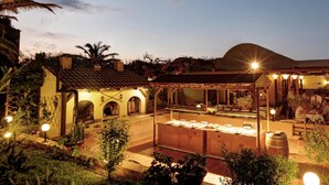 Un'area pranzo all'aperto con un lungo bancone, tavoli e sedie, circondata da piante e un edificio con tetto in tegole.