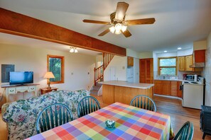 A cozy living room with a dining table, floral sofa, and a television. A kitchen with wooden cabinets and a microwave is visible in the background.