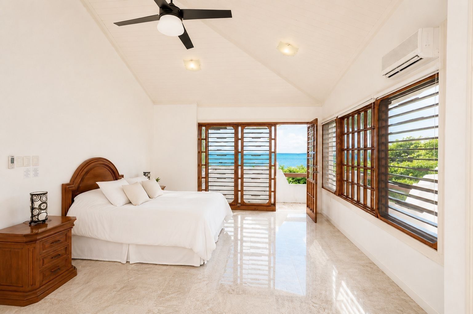 A bedroom with a bed, nightstand, ceiling fan, and large windows with a view of the ocean.