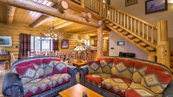 A log cabin interior with a wooden staircase, two sofas with patterned cushions, a chandelier, and a dining area.