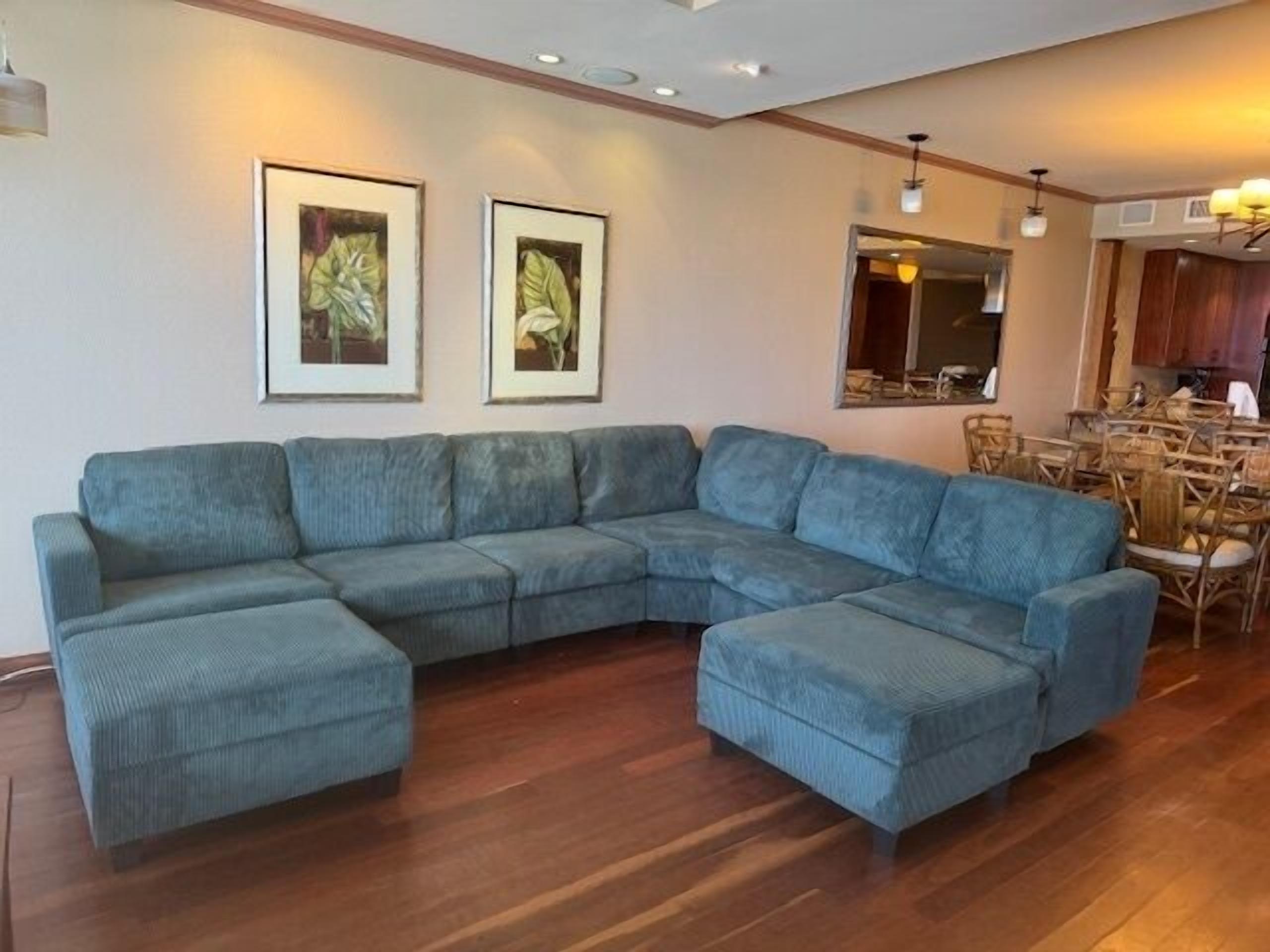 Ein Wohnzimmer mit Ecksofa, Holzboden, gerahmten Bildern an der Wand und einem Essbereich mit Stühlen und einem Tisch.