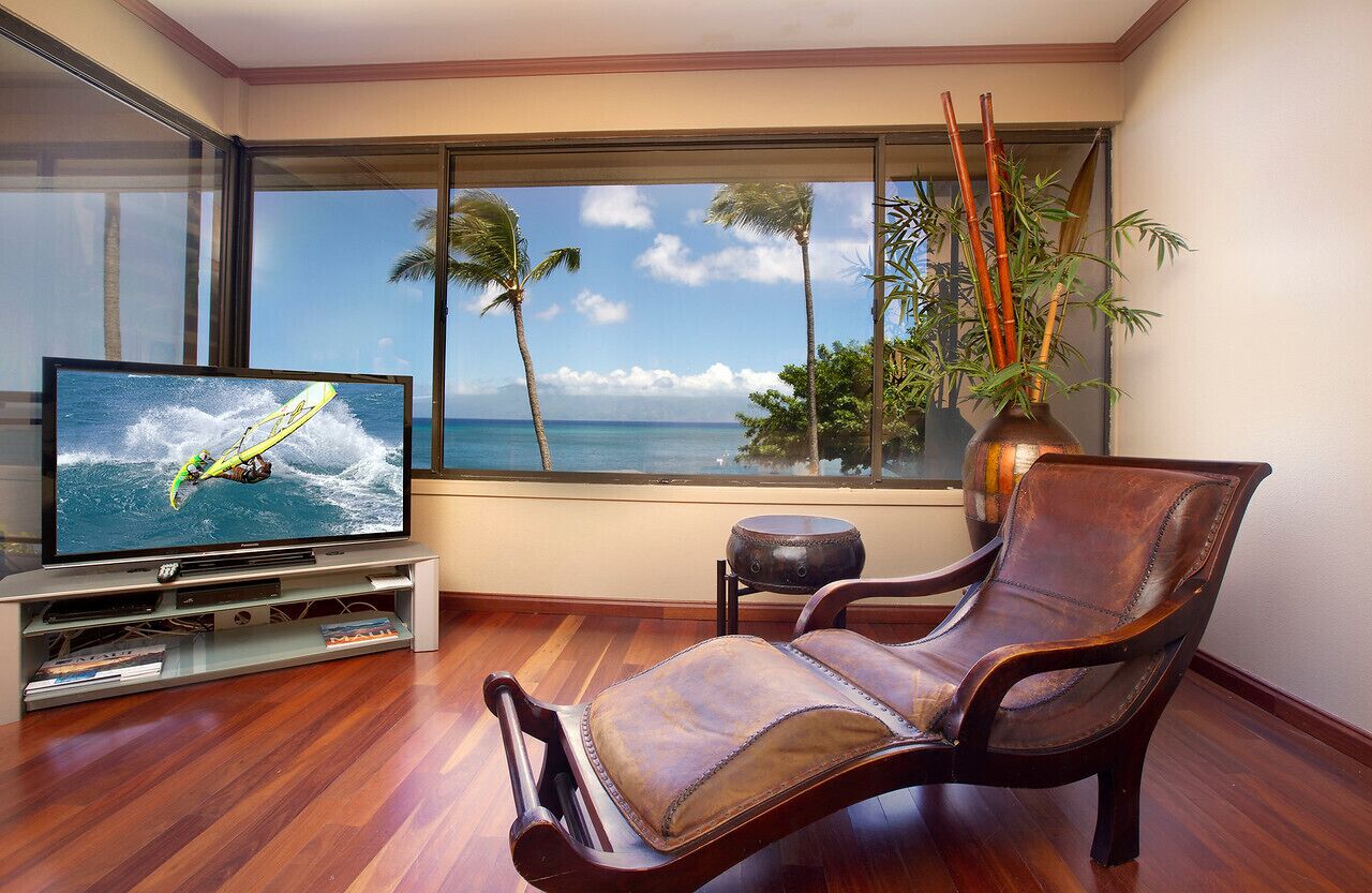 Una sala de estar con un televisor grande mostrando una escena de surf, un sillón de cuero, una mesita y vista al océano a través de amplios ventanales.