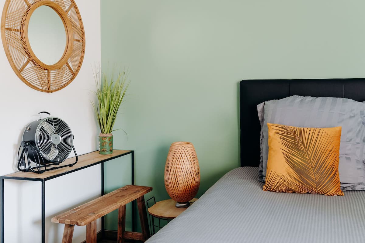 Une chambre à coucher avec un lit, une table de chevet, un ventilateur, un vase avec des plantes et un miroir au mur.
