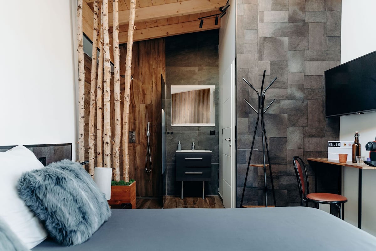 Une chambre à coucher moderne avec un lit, une armoire en bois, une salle de bain avec un lavabo et un miroir, et un téléviseur fixé au mur.