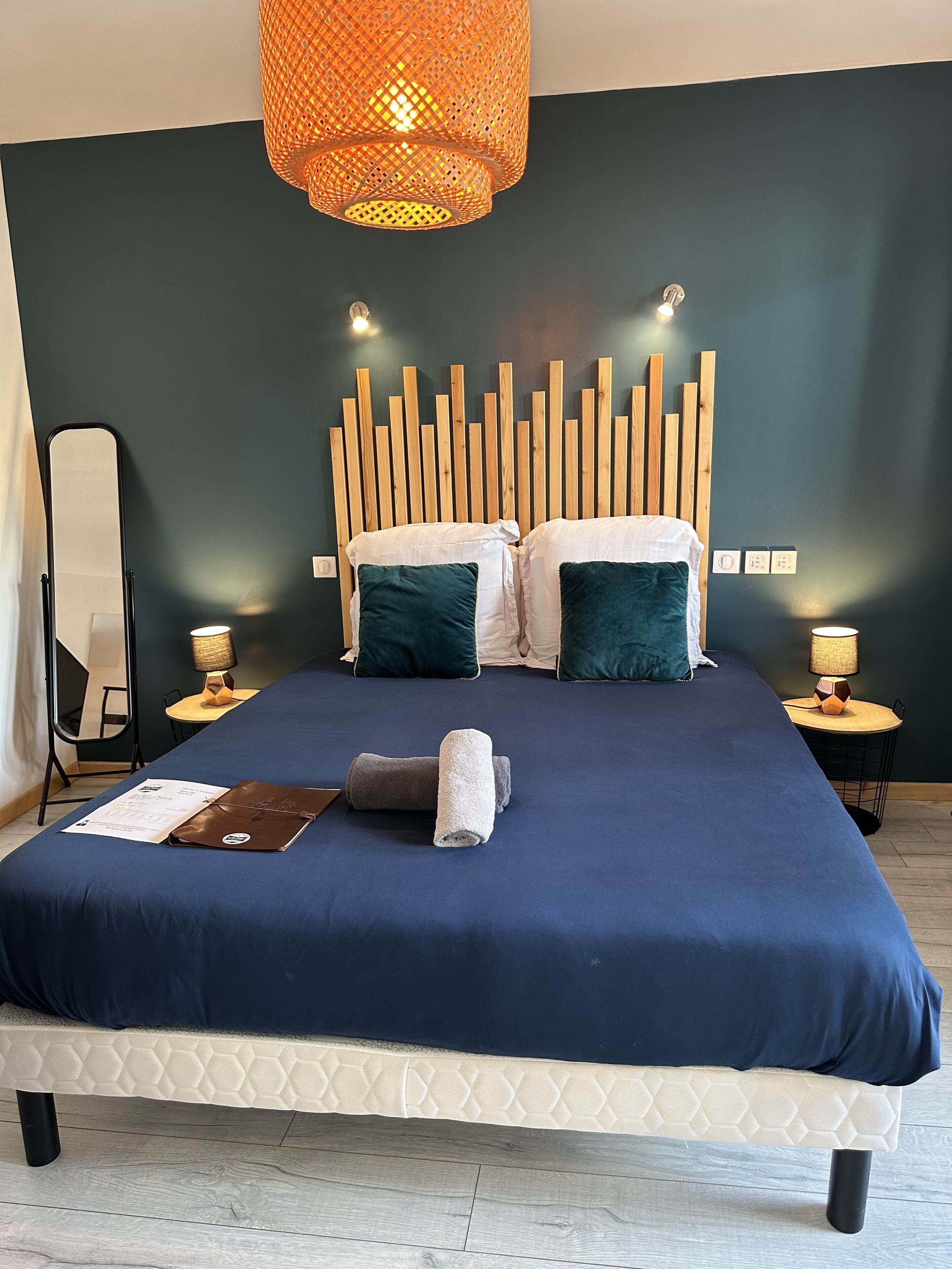 A neatly made bed with a blue bedspread, two pillows, and a rolled towel on a headboard with vertical wooden slats.