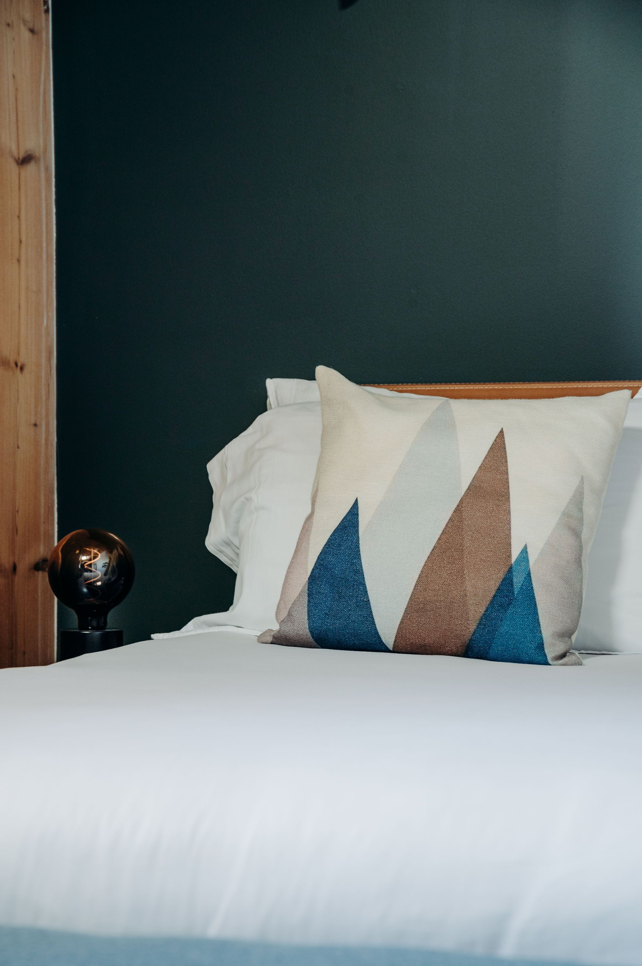 A bed with white bedding and a patterned pillow, a wooden nightstand with a lamp, and a dark green wall.