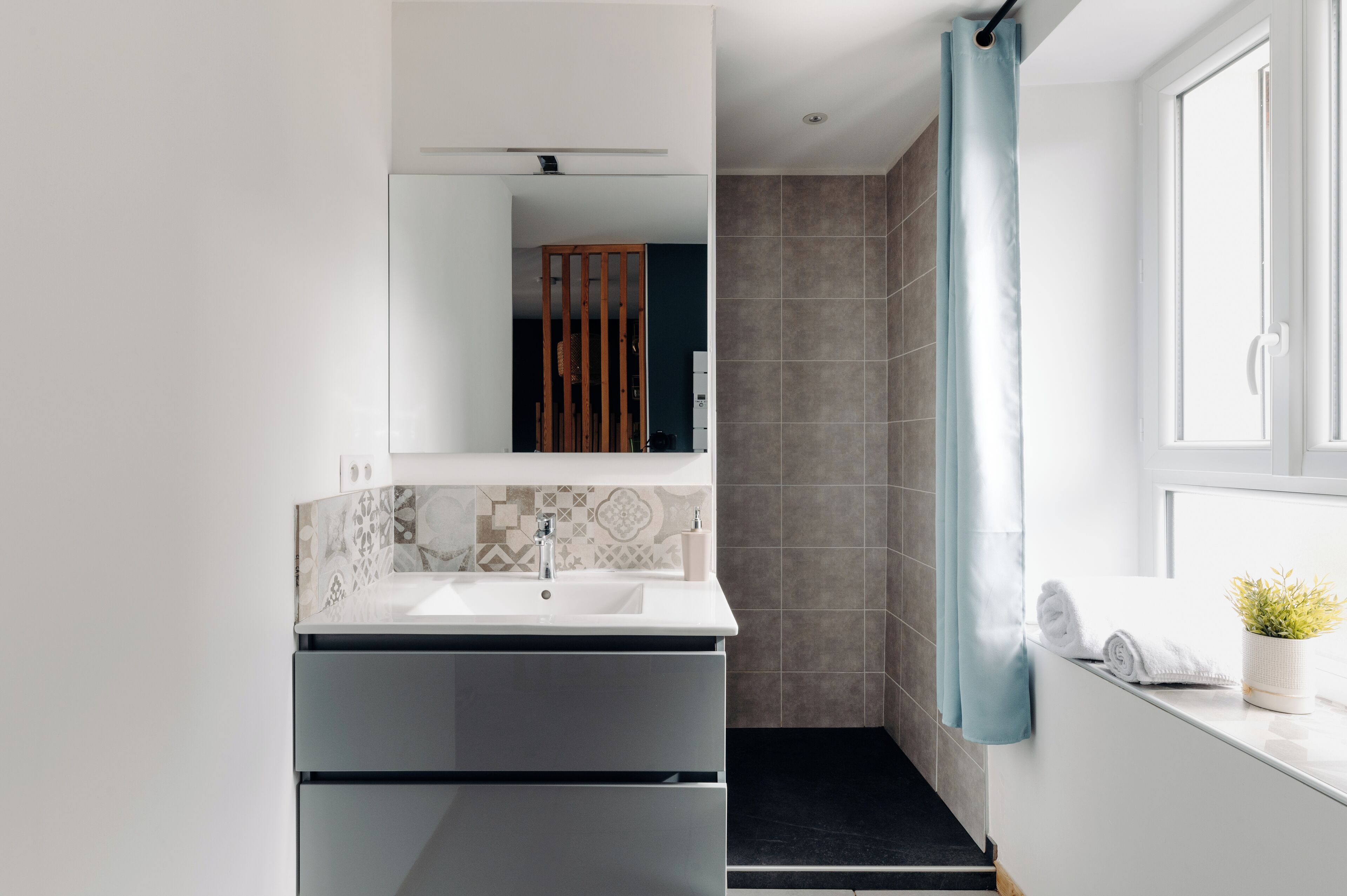A modern bathroom with a white sink, dark grey cabinets, a patterned tile backsplash, and a shower with a blue curtain.