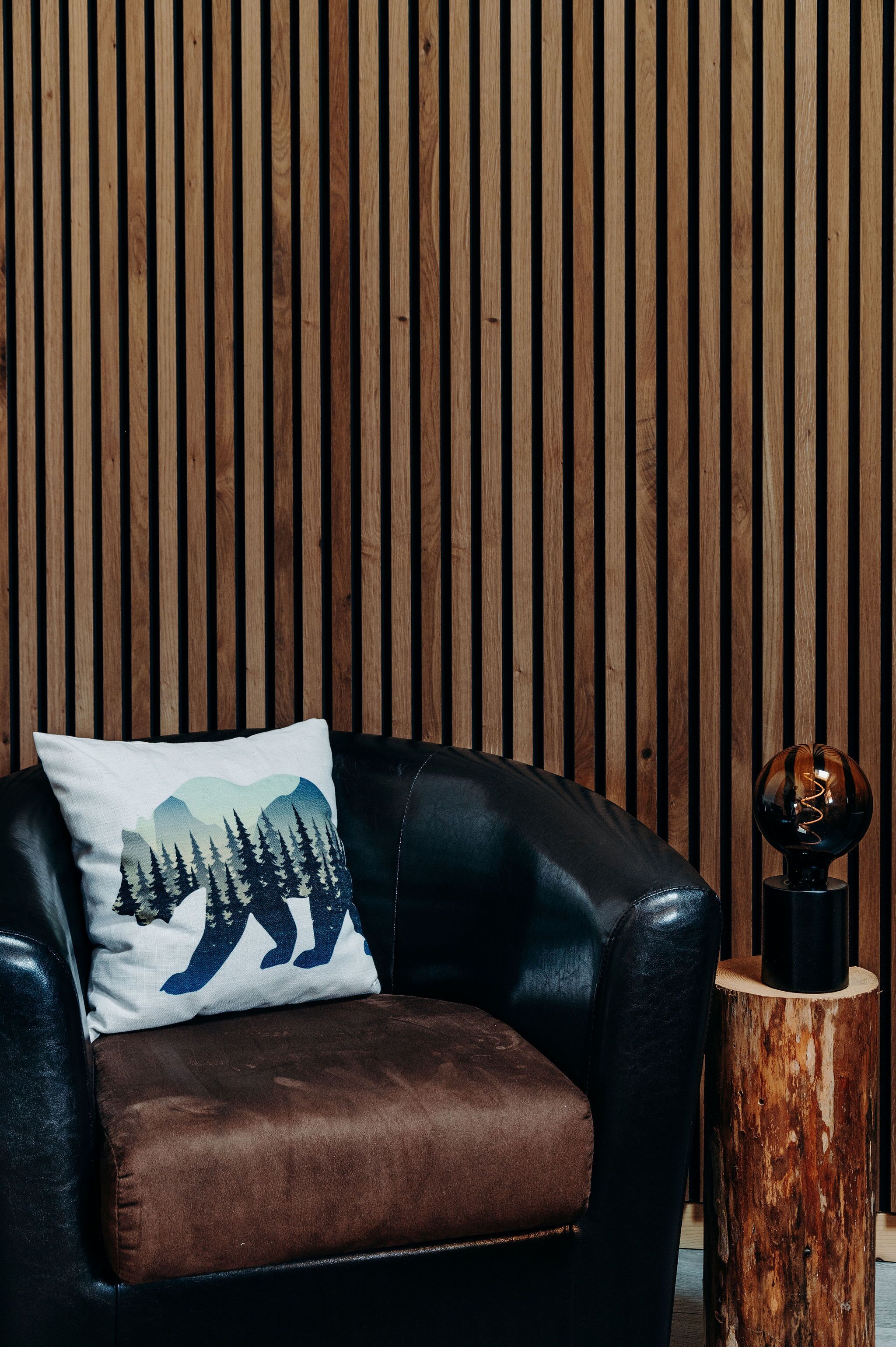 A leather armchair with a pillow featuring a mountain and bear design, positioned against a wooden slat wall.