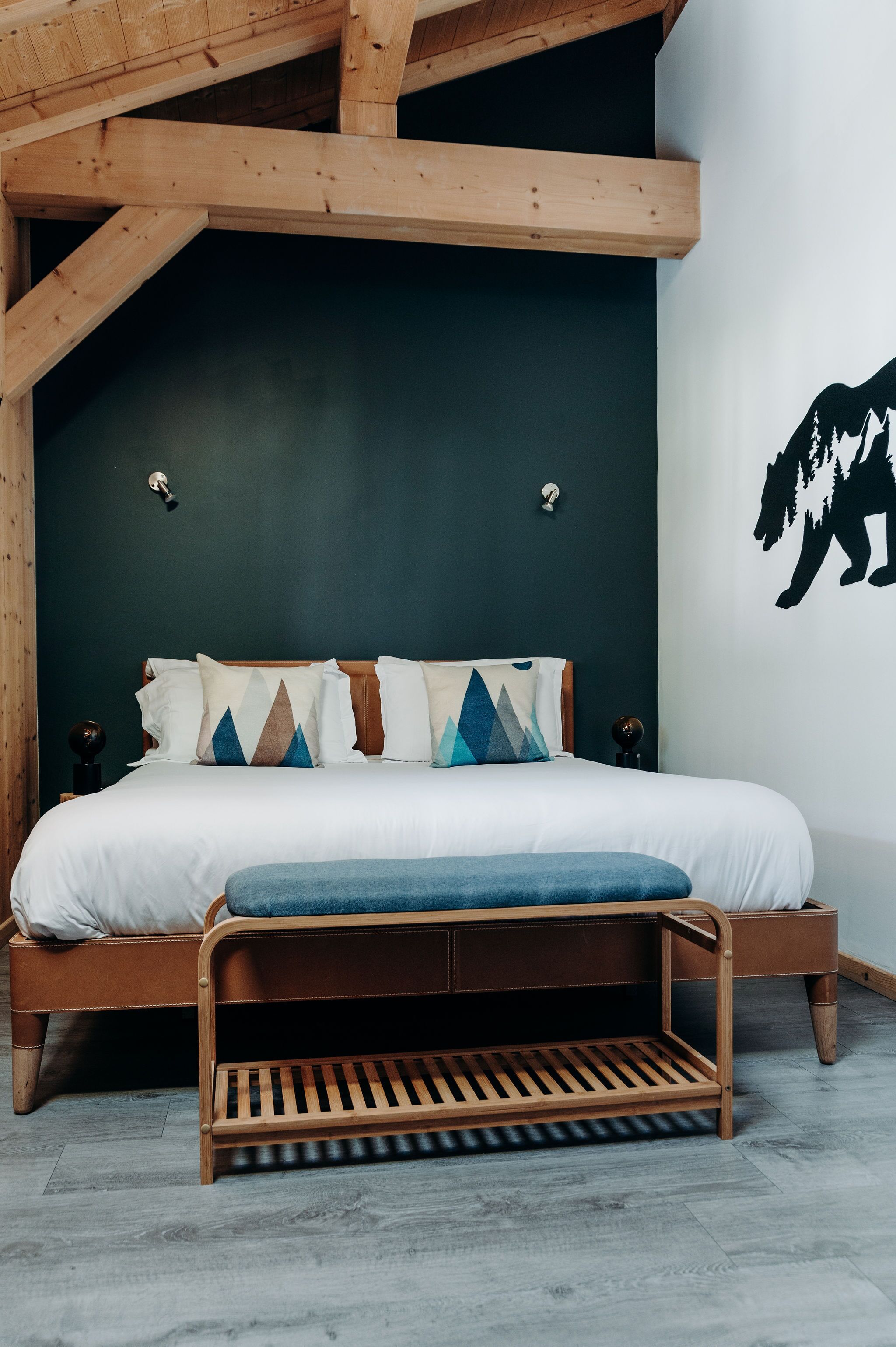 A bedroom with a wooden bed frame, a bench at the foot of the bed, and a wall with a black silhouette.