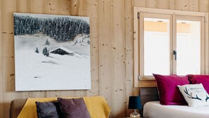 A cozy room with a wooden ceiling, a large painting of a snowy landscape, a yellow sofa with purple cushions, a round wooden table, and a bed with red and white pillows. - La Villa d'Hélène Chambres d’hôtes et Appartements-Cluses (Cluses)