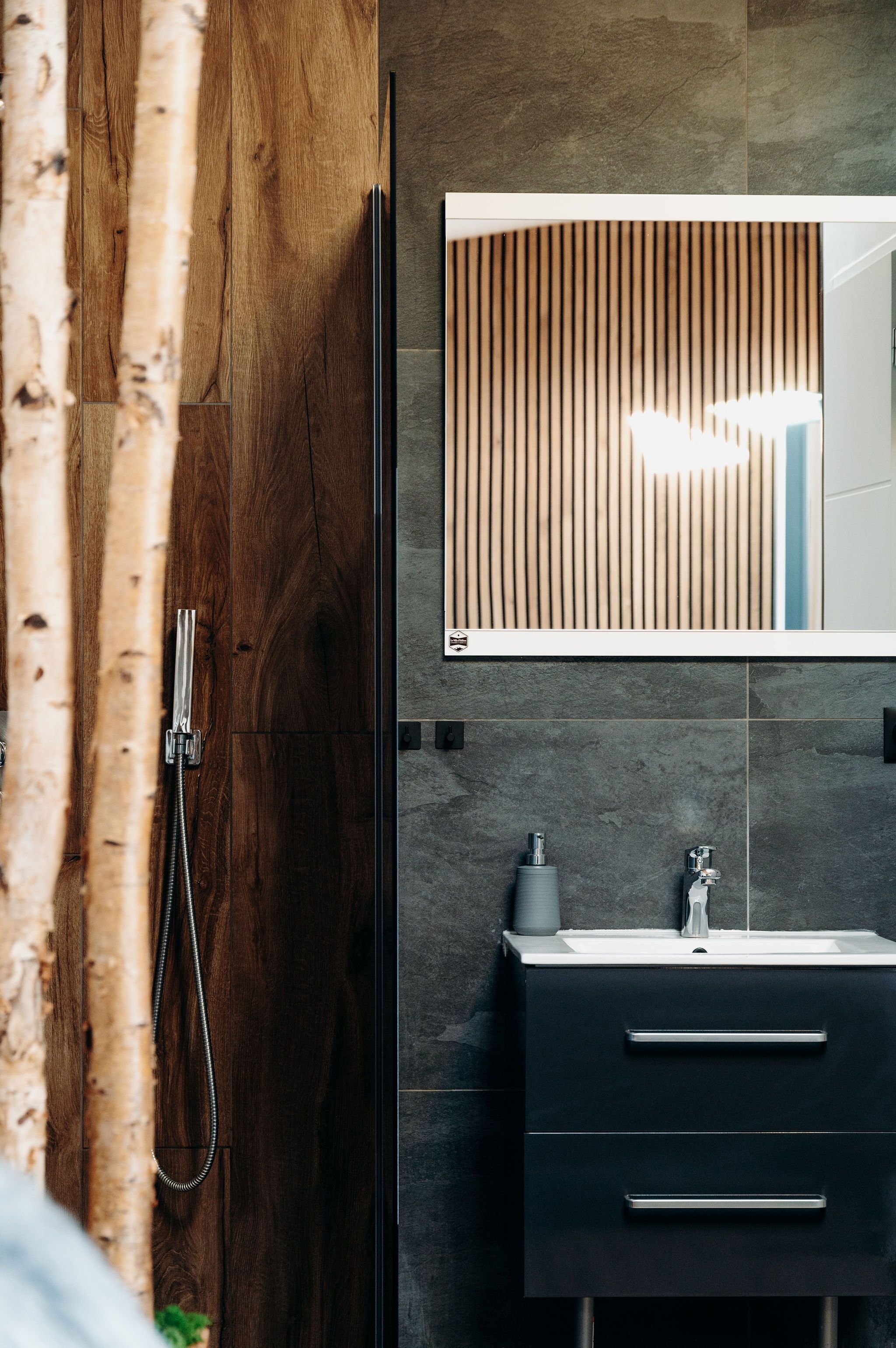 Un baño moderno con puerta de madera, ducha, espejo y tocador oscuro con lavabo.