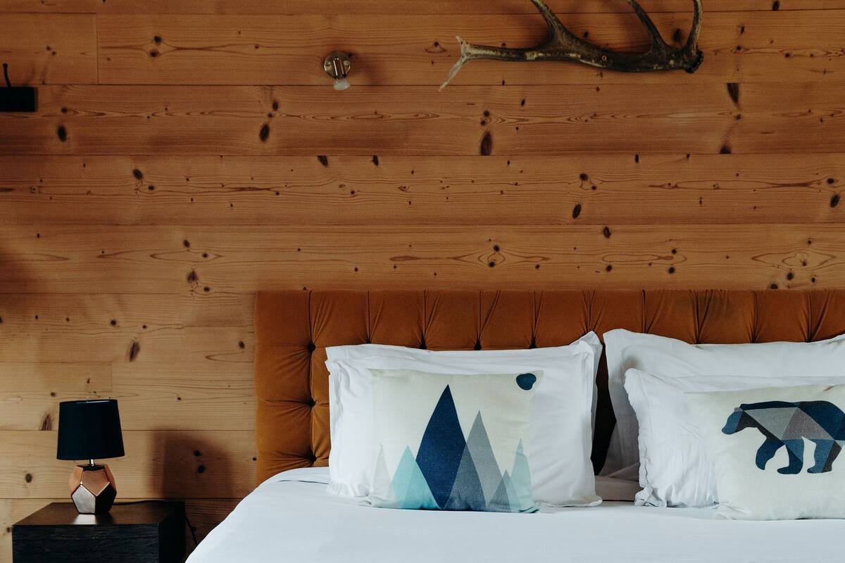 Une chambre à coucher avec une tête de lit en bois, un lit recouvert de linge de lit blanc, deux coussins décoratifs, une table de chevet avec une lampe et une paire de bois de cerf accrochée au mur.