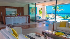 A modern living room with a white sofa, yellow cushions, and a coffee table. There is a kitchen area with white cabinets and a large window overlooking the ocean. - Luxury Villa Overlooking Orient Bay (SAINT MARTIN)