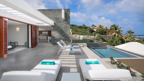 A modern outdoor pool area with white lounge chairs and a stone wall, overlooking a scenic landscape with palm trees and a view of the sea. - Luxury Villa Overlooking Orient Bay (SAINT MARTIN)