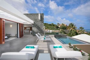 A modern outdoor pool area with white lounge chairs and a stone wall, overlooking a scenic landscape with palm trees and a view of the sea. - Luxury Villa Overlooking Orient Bay (SAINT MARTIN)