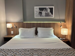 A hotel room with a bed, two bedside lamps, a framed picture on the wall, and a wooden headboard. - Promenade Soho Campos dos Goytacazes (Campos)