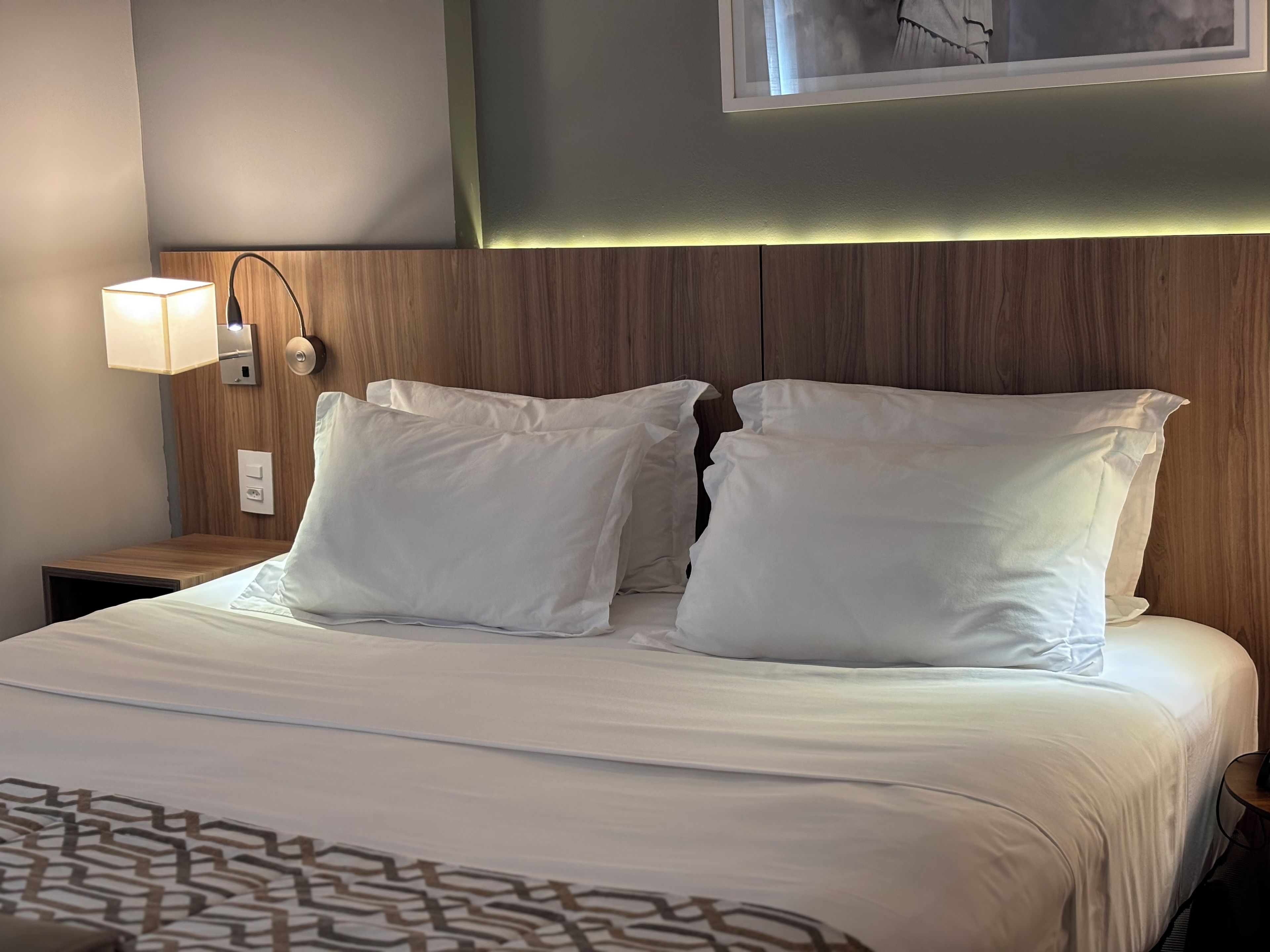 Una cama bien tendida con sábanas y almohadas blancas, una lámpara de noche y una lámpara de pared.